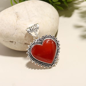 Puede incluir: Colgante en forma de coraz&oacute;n con una piedra roja oscura pulida en el centro. La piedra est&aacute; enmarcada por un engaste de metal plateado detallado con una fianza decorativa. Para un collar.