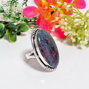 May include: A silver ring featuring a large, oval-shaped cabochon stone with a mottled pattern of deep magenta and blue-green hues. The metal setting is polished and decorated with a beaded texture along the outer edge of the stone bezel.