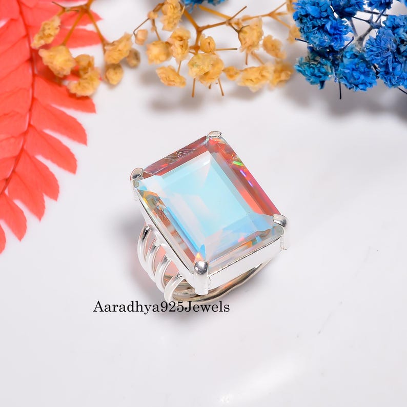 May include: A rectangular iridescent gemstone ring with a silver band. The gemstone reflects multiple colors, including blue, pink, and yellow. The ring is set against a white background with colorful floral accents. The text "Aaradhya925Jewels" is visible.