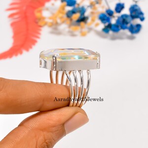 May include: A silver ring with a large, rectangular, iridescent gemstone. The ring has a multi-band shank and a wide, silver setting. The text "Aaradhya925Jewels" is visible on the ring.
