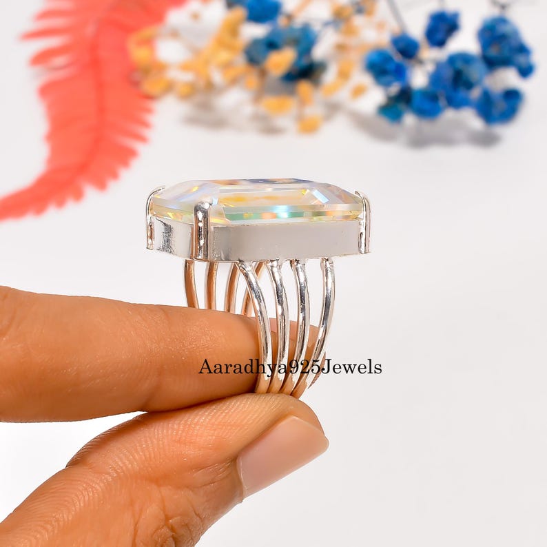 May include: A silver ring with a large, rectangular, iridescent gemstone. The ring has a multi-band shank and a wide, silver setting. The text "Aaradhya925Jewels" is visible on the ring.