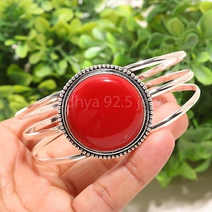 Peut inclure: Un bracelet manchette en argent avec une grosse pierre rouge vif et ronde au centre. La pierre est entourée d'une bordure argentée perlée. Le bracelet comporte trois bandes argentées incurvées. Le texte "dhya 92.5" est visible.