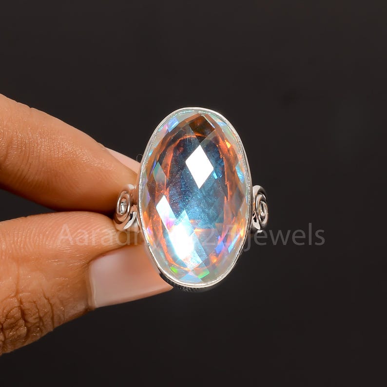 May include: A silver ring with a large, oval gemstone displaying iridescent colours. The faceted surface of the gemstone reflects light, producing a rainbow effect. The ring's band features a decorative swirl design on either side.