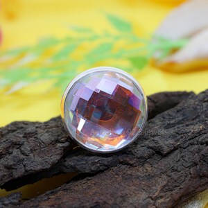May include: A round, faceted gemstone ring with a silver band. The gemstone has a clear, iridescent appearance, reflecting multiple colors. The ring is set on a piece of dark, textured wood against a yellow background.