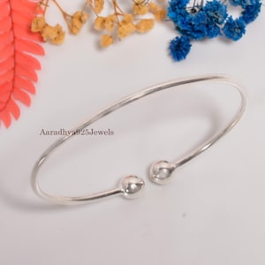 May include: A silver-colored open cuff bracelet with two ball-shaped ends. The bracelet is curved and has the text "Aaradhya925Jewels" on the left side. The background includes colorful flowers.
