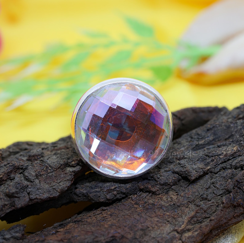 May include: A round, faceted gemstone ring with a silver band. The gemstone has a clear, iridescent appearance, reflecting multiple colors. The ring is set on a piece of dark, textured wood against a yellow background.