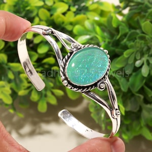 Puede incluir: Una pulsera de plata con una piedra preciosa ovalada verde iridiscente. La piedra está engastada en un bisel decorativo, y la pulsera tiene un diseño abierto. El texto "adh 925 jewels" es visible en la pulsera.