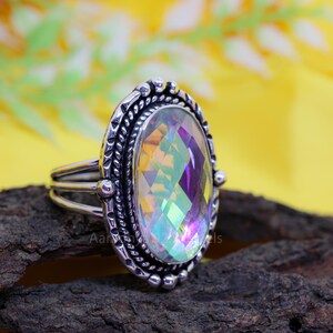 May include: A silver ring with an oval, iridescent gemstone. The gemstone displays a rainbow of colors and is set in a detailed, textured silver band. The ring has a triple-band design and is displayed on a dark, textured surface.
