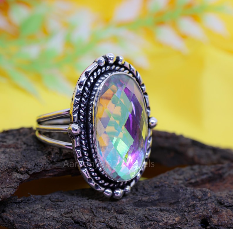 May include: A silver ring with an oval, iridescent gemstone. The gemstone displays a rainbow of colors and is set in a detailed, textured silver band. The ring has a triple-band design and is displayed on a dark, textured surface.