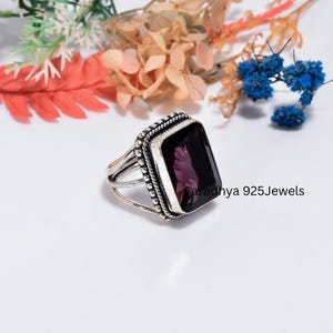 May include: A silver ring featuring a large, rectangular, deep purple gemstone set in a beaded metal frame. The band consists of three thin, parallel silver wires that converge at the base. The text Aaradhya 925Jewels is visible on the side.
