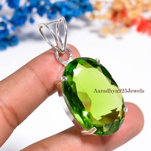 May include: A silver pendant featuring a large, oval-cut, lime-green gemstone. The gemstone is set in a silver bezel with a loop for a chain. The text "Aaradhya925Jewels" is visible on the right side of the pendant.