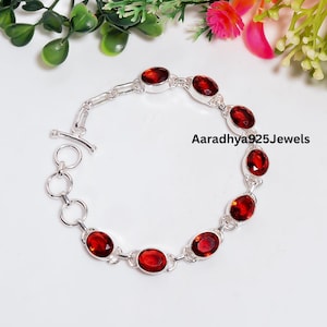 Op de afbeelding: Een zilveren kettingarmband met zeven ovaal geslepen, dieprode granaat edelstenen in individuele zettingen. Het armband heeft een knevelsluiting en een reeks cirkelvormige schakels. De tekst Aaradhya925Jewels is in het midden afgedrukt.