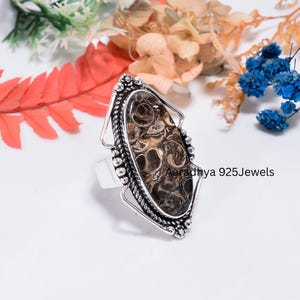 May include: A silver ring featuring an elongated oval fossil stone with brown and cream swirling patterns. The setting has a twisted rope border and decorative metal accents. The text Aaradhya 925Jewels is visible on the right side of the ring.