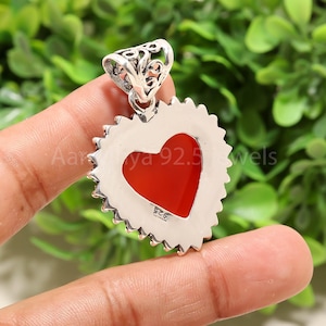 Puede incluir: Un colgante en forma de coraz&oacute;n con un centro rojo y un borde plateado texturizado. El colgante tiene una fianza decorativa y est&aacute; sostenido por los dedos de una persona. El texto "Aar ya 92.5 wels" es visible en el colgante.