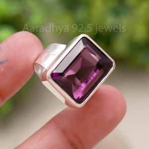 May include: A silver ring with a large, rectangular, deep purple gemstone. The ring has a simple, modern design with a wide band. The text "Aaradhya 92.5 jewels" is visible in the background.