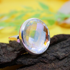 May include: A silver ring featuring a large, faceted, clear gemstone. The gemstone reflects a rainbow of colors. The ring is set on a dark, textured surface with a yellow background.