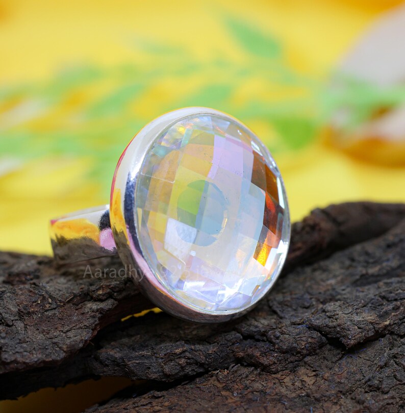 May include: A silver ring featuring a large, faceted, clear gemstone. The gemstone reflects a rainbow of colors. The ring is set on a dark, textured surface with a yellow background.