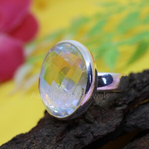 May include: A silver ring with a large, faceted, iridescent gemstone. The gemstone reflects a spectrum of colors, including yellow, orange, and blue. The ring is set on a dark, textured surface with the text "Aaradhya 925 Jewels".