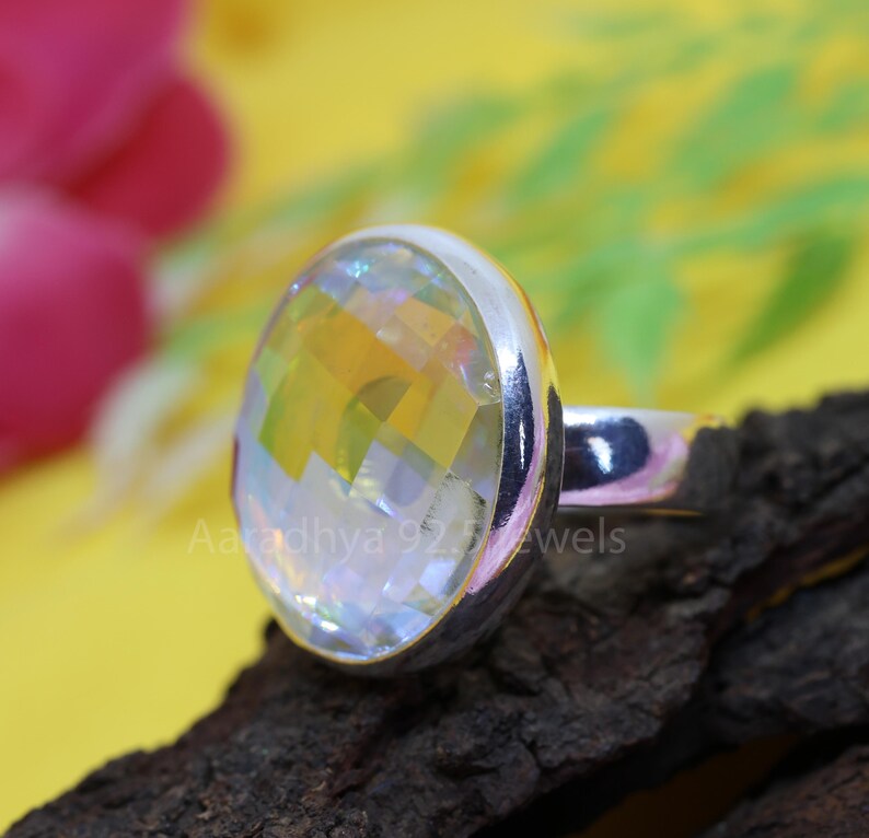 May include: A silver ring with a large, faceted, iridescent gemstone. The gemstone reflects a spectrum of colors, including yellow, orange, and blue. The ring is set on a dark, textured surface with the text "Aaradhya 925 Jewels".
