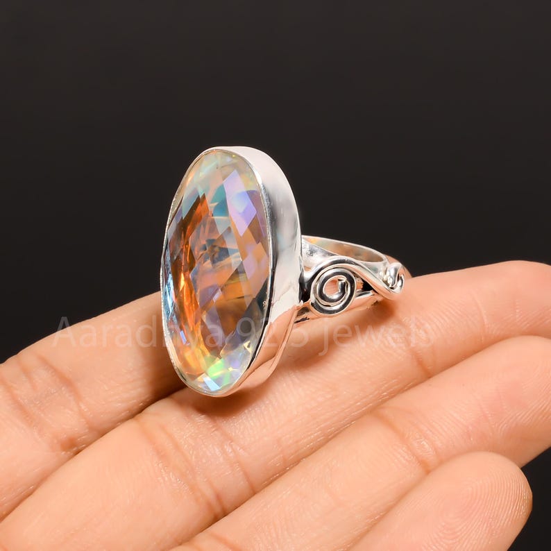 May include: A silver ring with a large, oval gemstone displaying iridescent colours. The gemstone has orange, yellow, and blue hues. The ring features a decorative swirl design on the band.