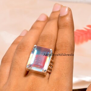 May include: A large, rectangular gemstone ring with a silver band. The gemstone has a rainbow iridescent effect, with colors of red, blue, and green. The text "Aaradhya925Jewels" is visible on the ring.