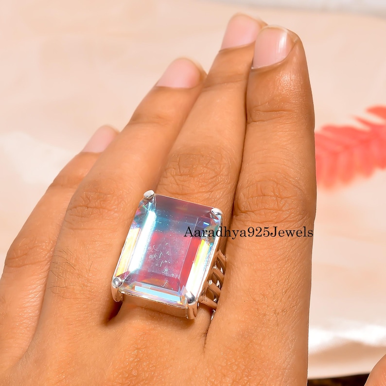 May include: A large, rectangular gemstone ring with a silver band. The gemstone has a rainbow iridescent effect, with colors of red, blue, and green. The text "Aaradhya925Jewels" is visible on the ring.