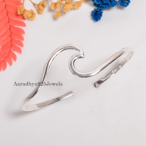 May include: A silver-colored cuff bracelet with a wave design. The open-ended bracelet features a curved wave shape in the center. The text "Aaradhya925Jewels" is visible on the bracelet. The bracelet is displayed on a white surface with colorful floral accents.