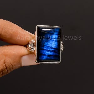 Puede incluir: Anillo de plata con una piedra preciosa azul rectangular e iridiscente. La piedra tiene una base azul oscuro con vetas azules más claras. El anillo tiene espirales decorativas y el texto "Aaradhya 925 jewels".