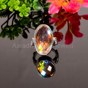 May include: A silver ring featuring a large, oval-cut, iridescent gemstone. The gemstone displays a spectrum of colours, including orange, yellow, and green. The ring has decorative swirls on the sides. The ring is shown on a reflective surface.