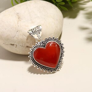 Puede incluir: Colgante en forma de coraz&oacute;n con una piedra roja oscura pulida en el centro. La piedra est&aacute; enmarcada por un engaste de metal plateado detallado con una fianza decorativa. Probablemente sea una pieza de joyer&iacute;a.