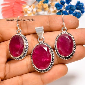 Op de afbeelding: Een sieradenset met een hanger en oorbellen, elk met ovale, gefacetteerde, dieprode edelstenen. De edelstenen zijn gezet in zilverkleurig metaal met een gedraaid touwontwerp. De tekst "Aaradhya925Jewels" is zichtbaar.