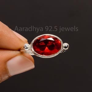 Può includere: Anello in argento con una grande gemma rossa scuro a taglio ovale. L'anello ha una fascia semplice con piccoli dettagli decorativi in argento. Il testo "Aaradhya 92.5 jewels" è visibile. La gemma è di un rosso intenso.