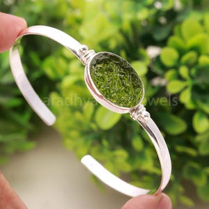 Puede incluir: Una pulsera de plata con una gema verde ovalada y texturizada. La pulsera tiene un diseño simple y elegante con un acabado pulido. La gema es el punto focal, engastada en un bisel de plata.