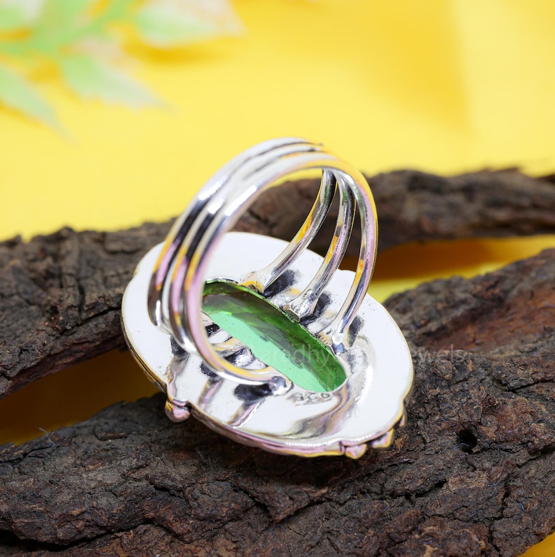 May include: A silver ring with a large, oval-shaped, light green gemstone. The ring has a triple-band design and a detailed setting. The gemstone is set in a silver bezel, and the ring is displayed on a piece of dark wood.