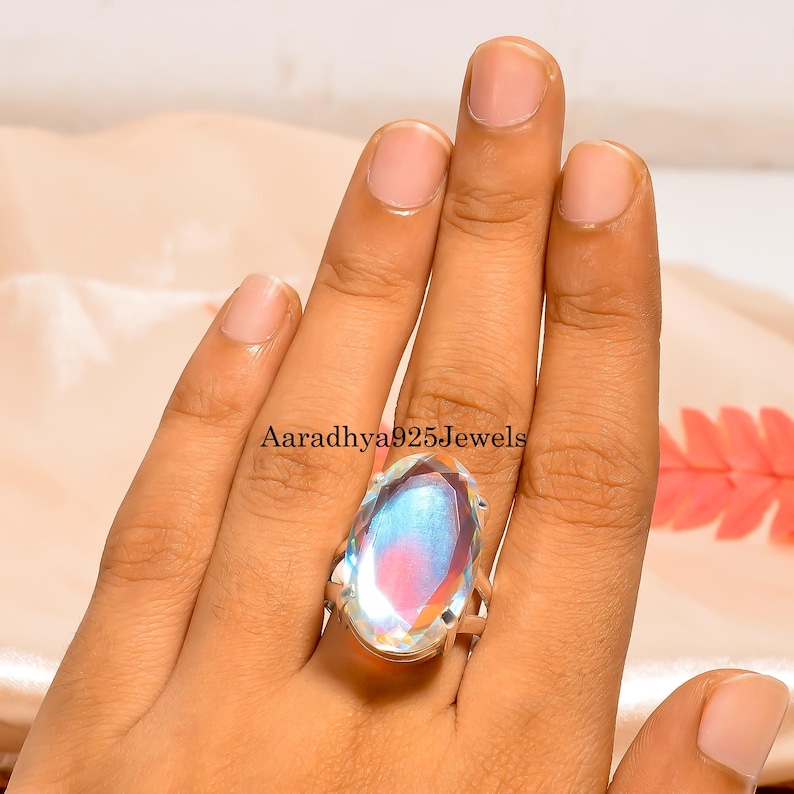 May include: A large, oval-cut gemstone ring with a silver band. The gemstone displays a rainbow of colors, including blue, pink, and yellow. The ring is set on a hand, showcasing the size and brilliance of the stone. The text "Aaradhya925Jewels" is visible.
