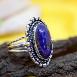 May include: A silver ring with an oval, deep blue gemstone. The gemstone is set in a decorative silver bezel with a textured design. The ring band has a triple-band design. The text "aradhya 92.5 jewels" is visible.