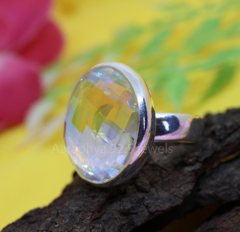 May include: A silver ring with a large, faceted, iridescent gemstone. The gemstone reflects a spectrum of colors, including yellow, orange, and blue. The ring is set on a dark, textured surface with the text "Aaradhya 925 Jewels".