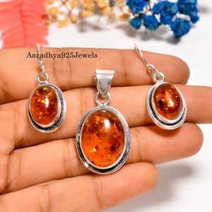 Può includere: Un set di gioielli in argento con ambra. Il set comprende un ciondolo e orecchini, ciascuno con una pietra d'ambra ovale. L'ambra ha un colore arancione-marrone caldo. Il marchio "Aaradhya925Jewels" è visibile.