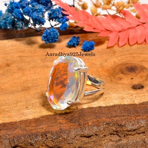 May include: A silver ring with a large, faceted, oval-cut gemstone. The gemstone has a rainbow iridescence, with colors of orange, yellow, and green. The ring is set on a wooden surface, with blue and red floral accents in the background. The text "Aaradhya925Jewels" is visible.