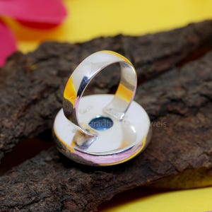 May include: A silver ring with a large, round, light blue gemstone. The ring band is thick and polished, and the gemstone is set in a bezel setting. The ring is displayed on a piece of dark wood.