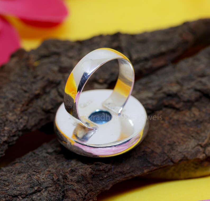 May include: A silver ring with a large, round, light blue gemstone. The ring band is thick and polished, and the gemstone is set in a bezel setting. The ring is displayed on a piece of dark wood.