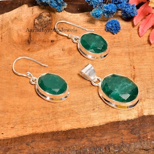Può includere: Un set di gioielli con un ciondolo e orecchini con pietre preziose ovali verde smeraldo incastonate in argento. Gli orecchini sono a gancio e il ciondolo ha un'attaccatura in argento. I gioielli sono esposti su una superficie di legno.