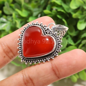 Puede incluir: Colgante en forma de coraz&oacute;n con una piedra roja vibrante engastada en un marco plateado. El marco tiene un borde de cuentas y una fianza decorativa. El texto "Adhya 92.5 Jewels" es visible en el colgante.