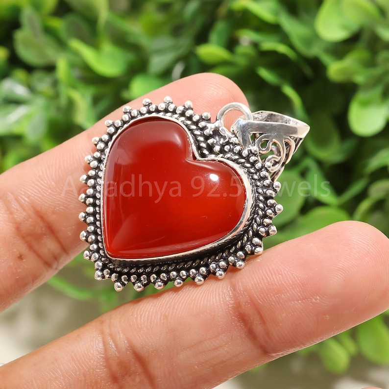 Puede incluir: Colgante en forma de coraz&oacute;n con una piedra roja vibrante engastada en un marco plateado. El marco tiene un borde de cuentas y una fianza decorativa. El texto "Adhya 92.5 Jewels" es visible en el colgante.