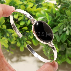 Könnte beinhalten: Ein silbernes Armreif-Armband mit einem großen, ovalen, schwarzen Onyxstein in der Mitte. Das Armband wird vor grünem Laub gehalten. Der Text "Asadhya 92.5 jewels" ist auf dem Armband zu sehen.
