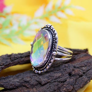 May include: A silver ring with an oval, iridescent gemstone. The gemstone displays a spectrum of colors, including green, yellow, and purple. The ring has a detailed, textured setting with a braided border and small bead accents.