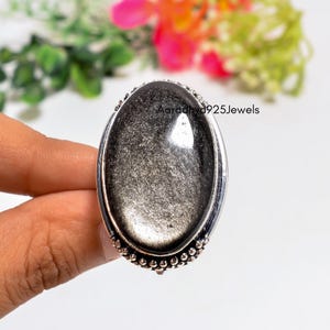 May include: An oval-shaped ring featuring a shimmering black and silver obsidian gemstone set in a polished silver band with beaded detailing along the edge. The text Aaradhya925Jewels is visible on the surface of the stone in this jewelry piece.