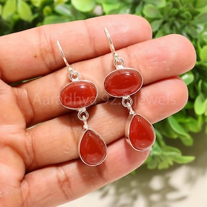 Può includere: Orecchini pendenti in argento con pietre rosse ovali e a goccia. Gli orecchini sono sospesi a ganci d'argento. Il testo "Aaradhya 92 jewels" è visibile. Lo sfondo è verde.