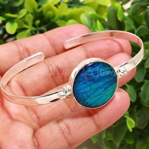 Op de afbeelding: Een zilveren manchetarmband met een ronde, iriserende blauw-groene middensteen. De armband heeft een eenvoudig, elegant ontwerp met een gepolijste afwerking. De middensteen heeft een unieke, getextureerde uitstraling. De armband wordt op een hand getoond.