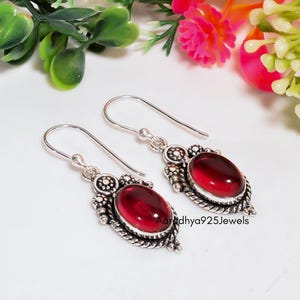 Peut inclure: Une paire de boucles d'oreilles pendantes en argent avec des pierres cabochons rouge foncé de forme ovale, serties dans des montures en métal orné avec des détails en corde torsadée et de petites sphères. Le texte Aaradhya925Jewels est visible.